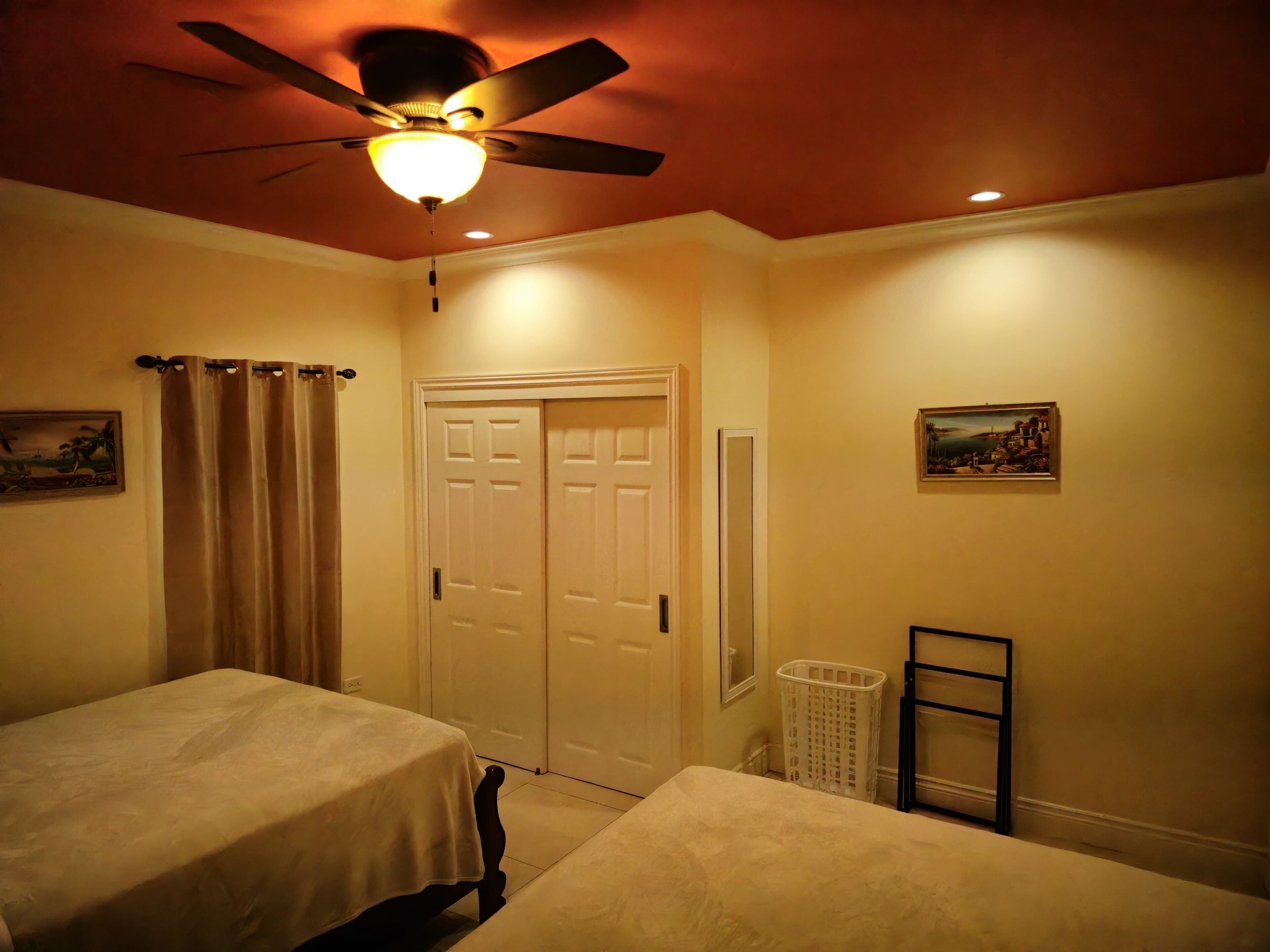 A bedroom with two beds, a ceiling fan, a door, a wall-mounted mirror, and a painting.