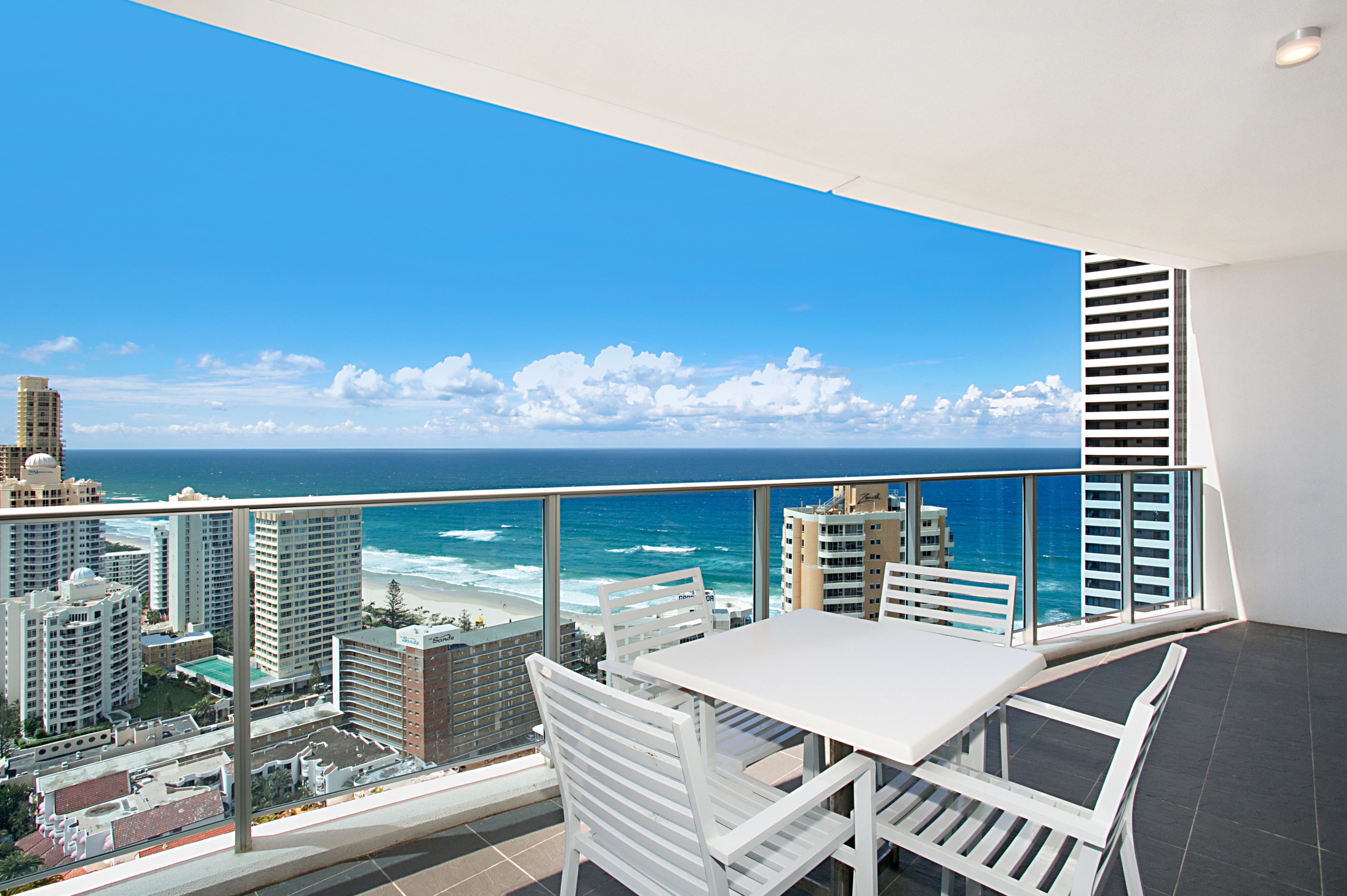 A balcony with a white table and chairs, overlooking a beach and ocean with buildings in the background.