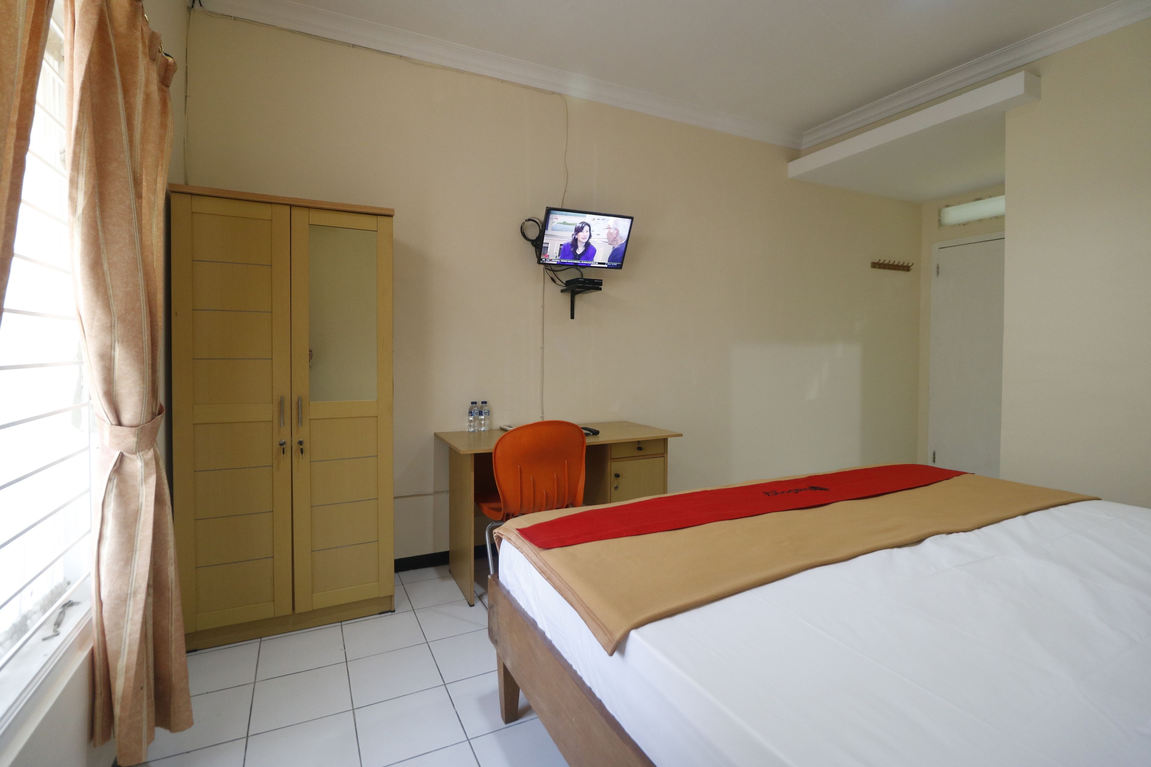 A hotel room with a bed, a desk with a chair, a television mounted on the wall, and a wardrobe.