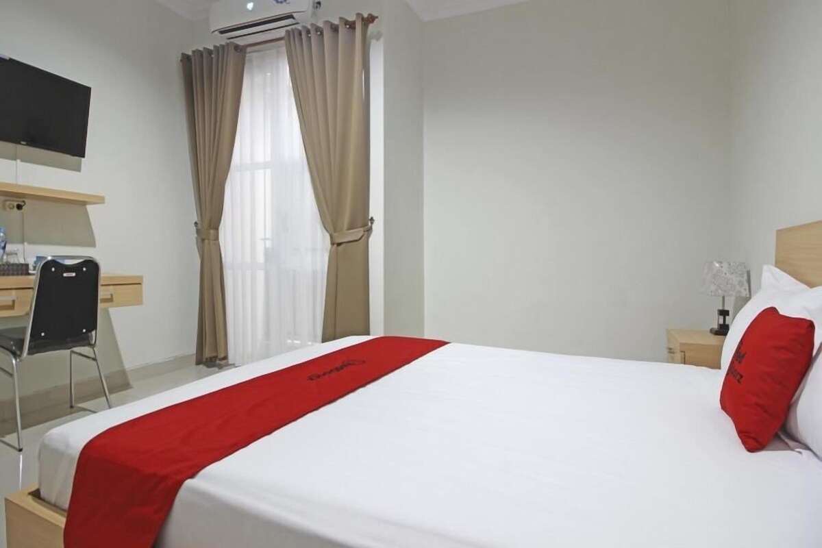 Una habitación de hotel con cama, escritorio, silla, televisión y ventana con cortinas.