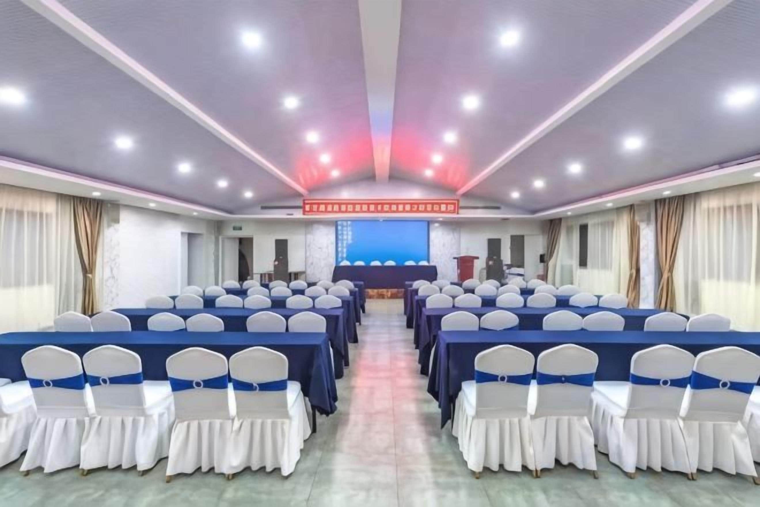 A conference room with rows of tables and chairs, a stage with a podium, and a large screen at the front.