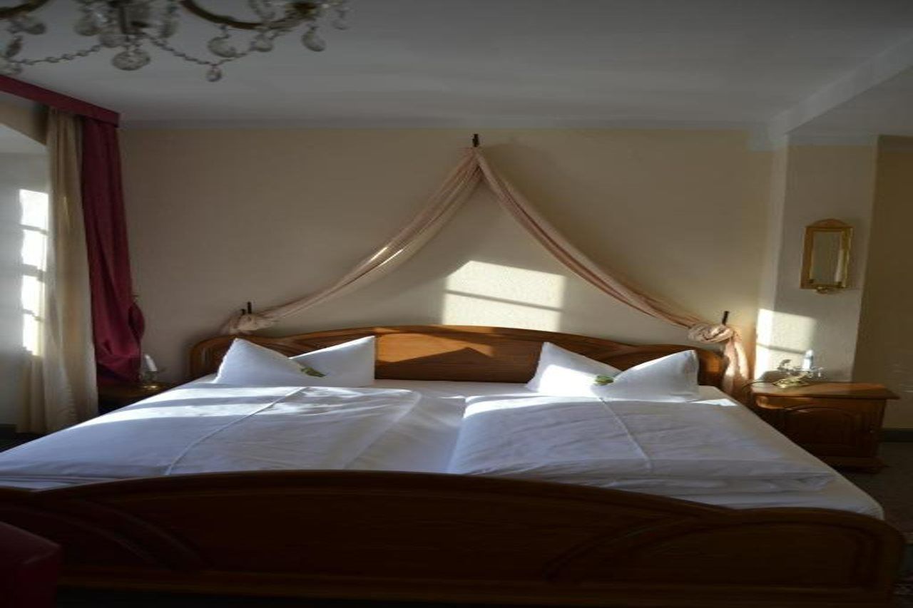 Ein Doppelbett mit Holz-Kopfteil, ein Nachttisch mit Lampe sowie ein Fenster mit Vorhängen.