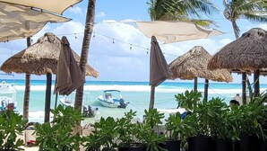 A beachside patio with thatched umbrellas, potted plants, and a view of the ocean and a boat. - Ocean view Condo, Large Private balcony over beach with jacuzzi, 2 pools, W/D (Playa del Carmen)
