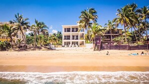 A beachfront property with a two-story building, palm trees, and a sandy beach. - "Seas the Day" at Sandy Beach (Rincon)