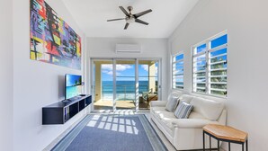 A modern living room with a large TV, a ceiling fan, and a view of the ocean through large windows. - "Seas the Day" at Sandy Beach (Rincon)