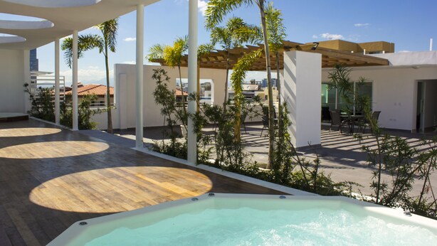 A rooftop pool area with a hot tub, wooden decking, palm trees, and a pergola.