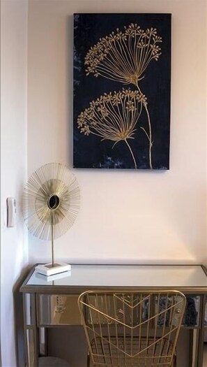 A room with a dark blue wall, a gold-framed picture of flowers, a glass table with a book, and a gold-framed chair.