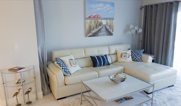 A living room with a beige sectional sofa, a glass coffee table, and a painting of a beach scene.