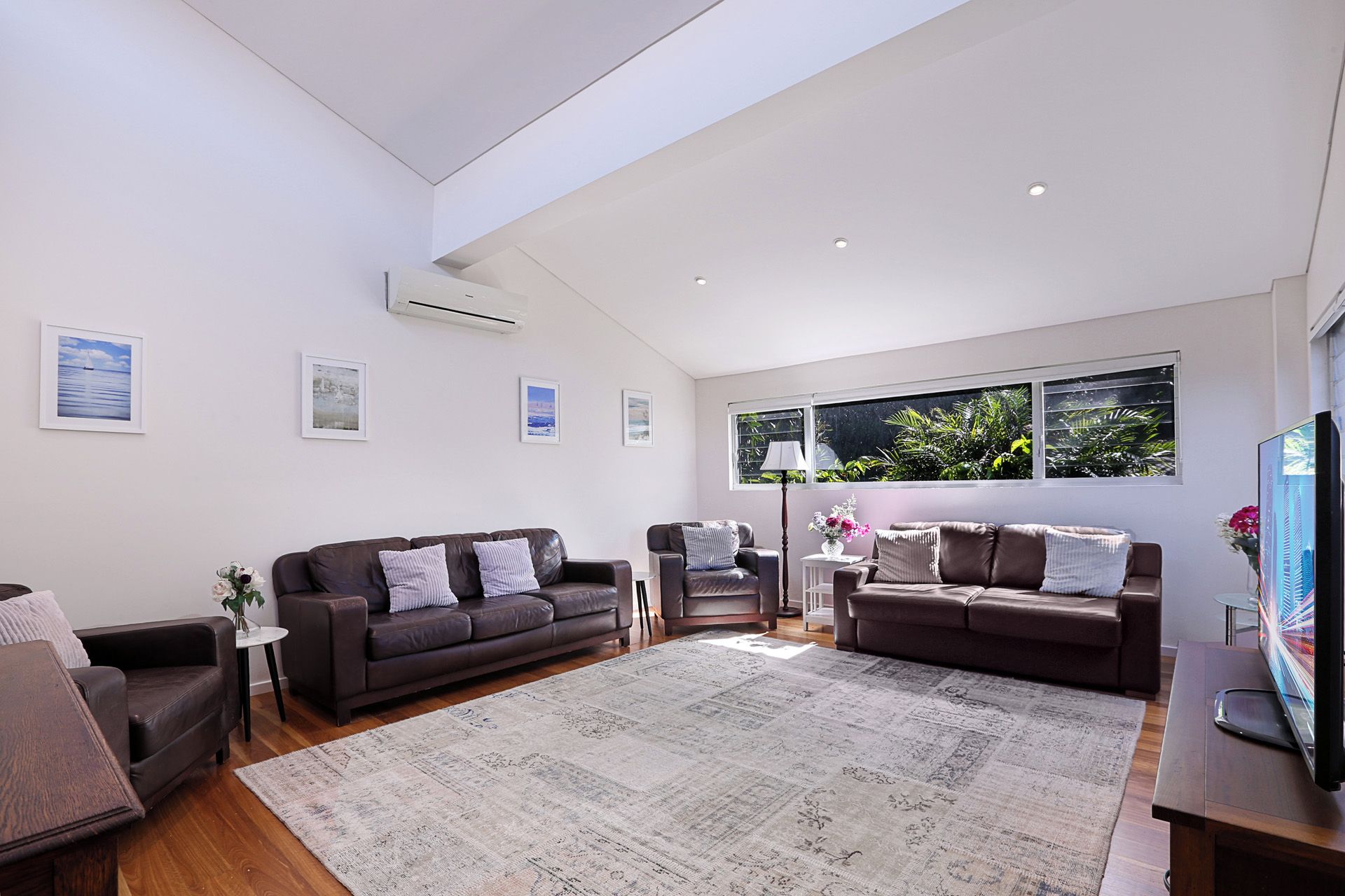 A modern living room with two sofas, a coffee table, and a TV. There are framed pictures on the wall and a window with a view of greenery.