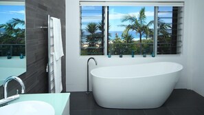Une salle de bain moderne avec une baignoire sur pieds, une douche à parois de verre et une grande fenêtre offrant une vue sur les palmiers et l’océan.