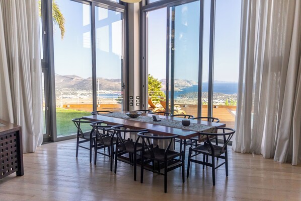 Un espace repas avec une longue table et des chaises, de grandes fenêtres donnant sur un paysage.