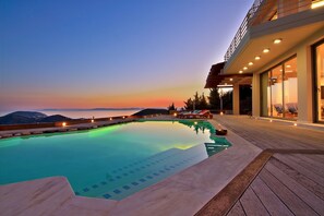 Une piscine avec vue sur les montagnes au crépuscule, une terrasse en bois et un bâtiment moderne doté de grandes fenêtres.