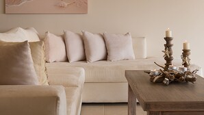 A living room with a beach-themed painting, a white sofa, a wooden coffee table with candles, and a wall-mounted light fixture. - Villa Gold in Petrothalassa (Porto Cheli)