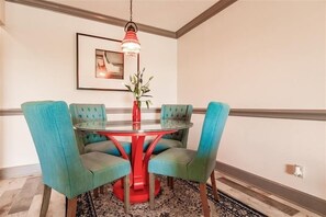A dining area with a round glass table, four teal upholstered chairs, a red base, a hanging red and white lamp, a framed picture on the wall, and a patterned rug on the floor. - Beachcomber unit 303 (Longboat Key)