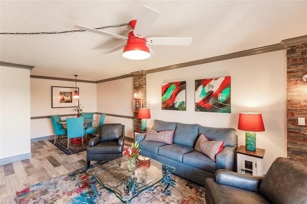 A living room with a blue sofa, two armchairs, a glass coffee table, and a red ceiling fan. There are two wall-mounted lamps and a framed picture on the wall. - Beachcomber unit 303 (Longboat Key)
