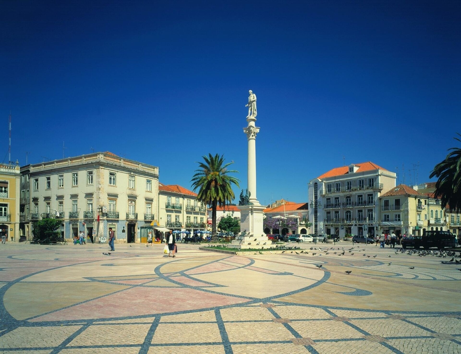 Una plaza pública con un monumento central, rodeada de edificios históricos con tejados de tejas rojas.