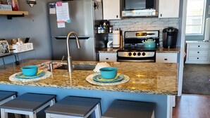 A modern kitchen with a granite countertop, stainless steel appliances, and bar stools. - NEW! Beach Chic-Pool-triple bunks-Slps 7-beach chairs & cart-elev-off BW (North Wildwood)