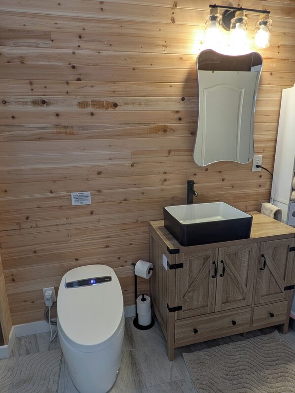 Une salle de bain avec des murs recouverts de panneaux en bois, des toilettes blanches, un meuble en bois avec des tiroirs, un miroir et un luminaire placé au-dessus du miroir.