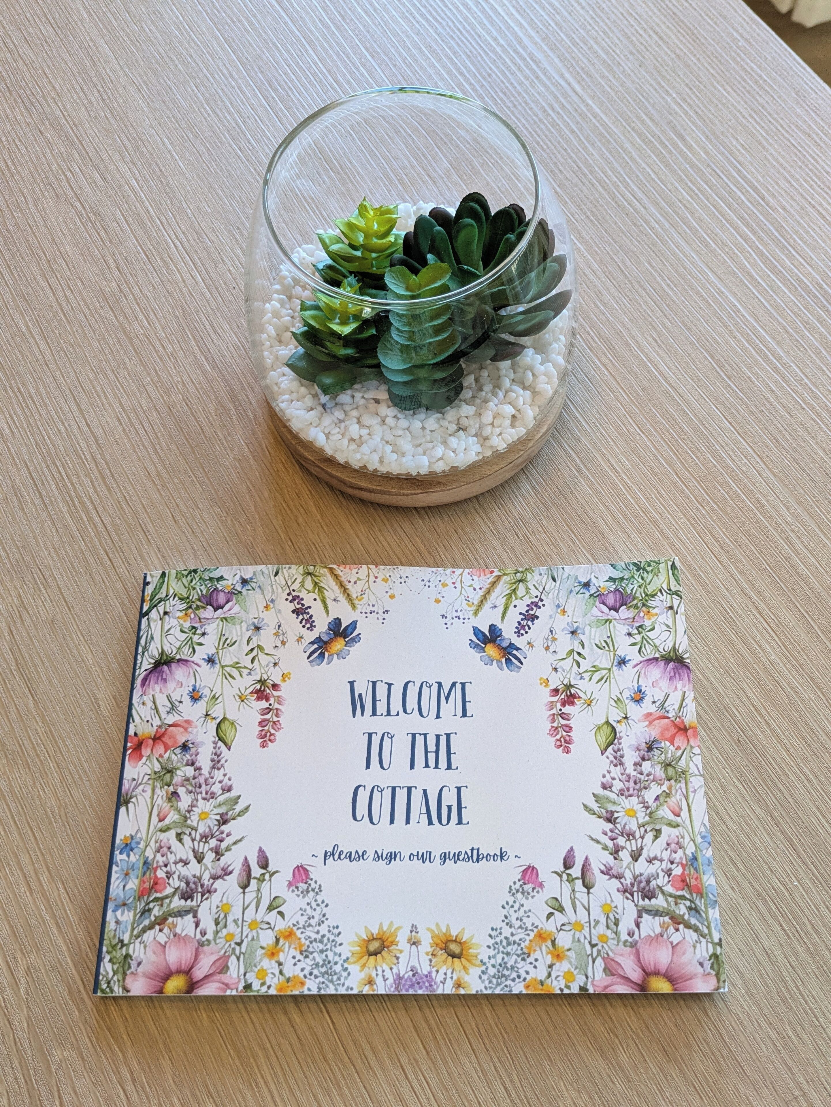 A glass terrarium with succulents and white pebbles on a wooden surface, next to a 'Welcome to the Cottage' sign with floral designs.