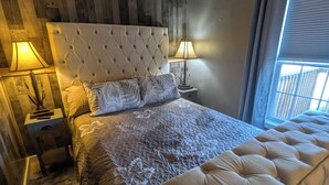 Un dormitorio con cabecera capitonada, una cama grande con colcha estampada, dos lámparas de noche, una alfombra mullida y vistas al exterior a través de una ventana.