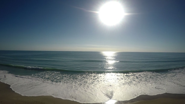 Una playa con olas, cielo despejado y el sol reflejándose en el agua.