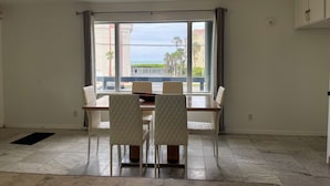 Un comedor con mesa y sillas, ventanas amplias con vistas a palmeras y un edificio, y una lámpara en el techo.