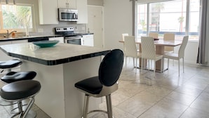 Una cocina moderna con una isla central, taburetes de bar, una zona de comedor con mesa y sillas, y un ventanal que ofrece vistas al exterior.