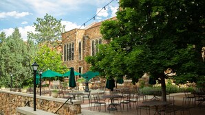 An outdoor patio with tables and chairs, a stone wall, and a building with arched windows in the background. - Glen Eyrie Castle & Conference Center A Ministry of the Navigators (Colorado Springs)