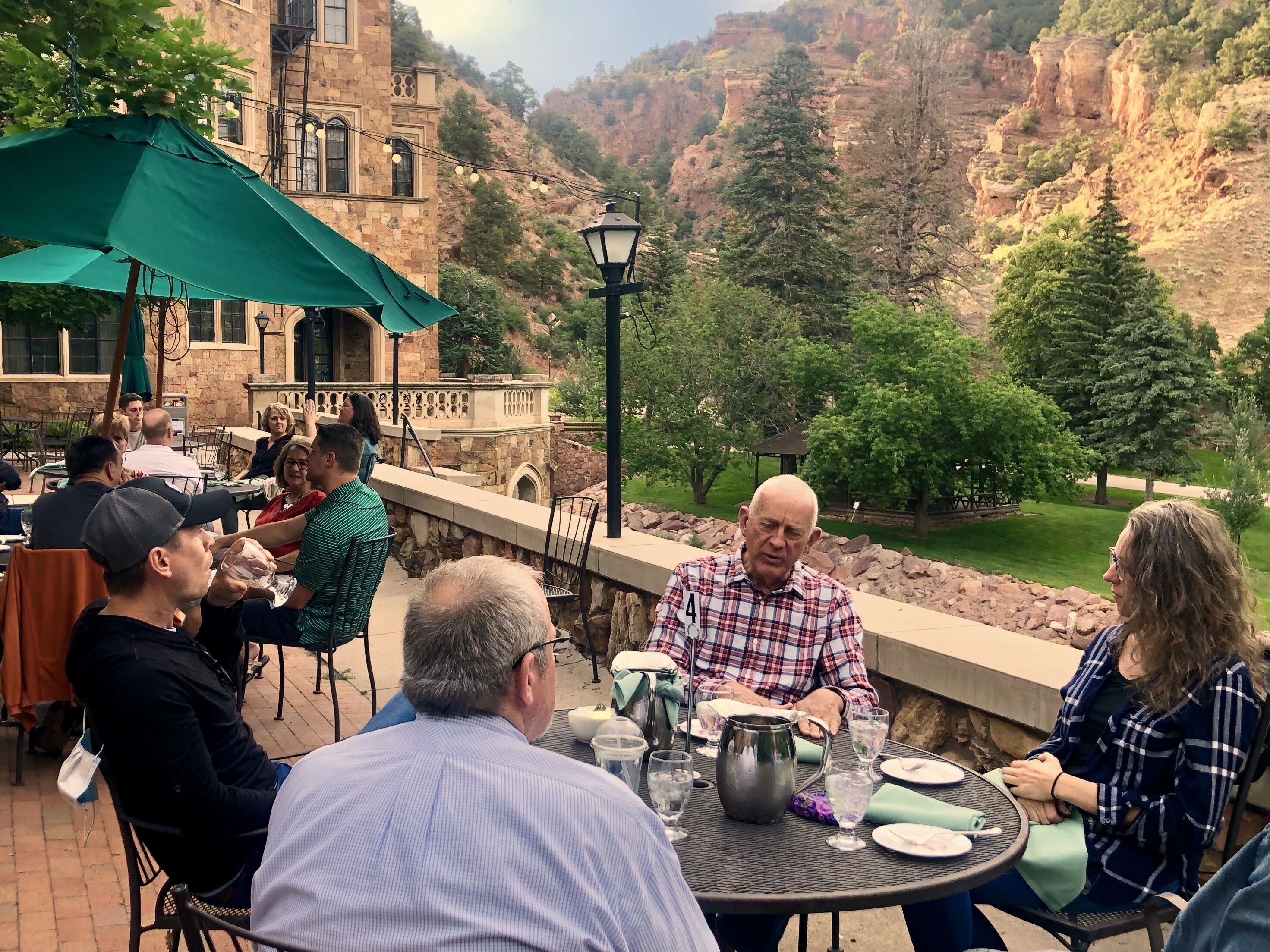 Un groupe de personnes est assis à des tables installées à l’extérieur, sous des parasols, et profite d’un repas tout en admirant un paysage montagneux.