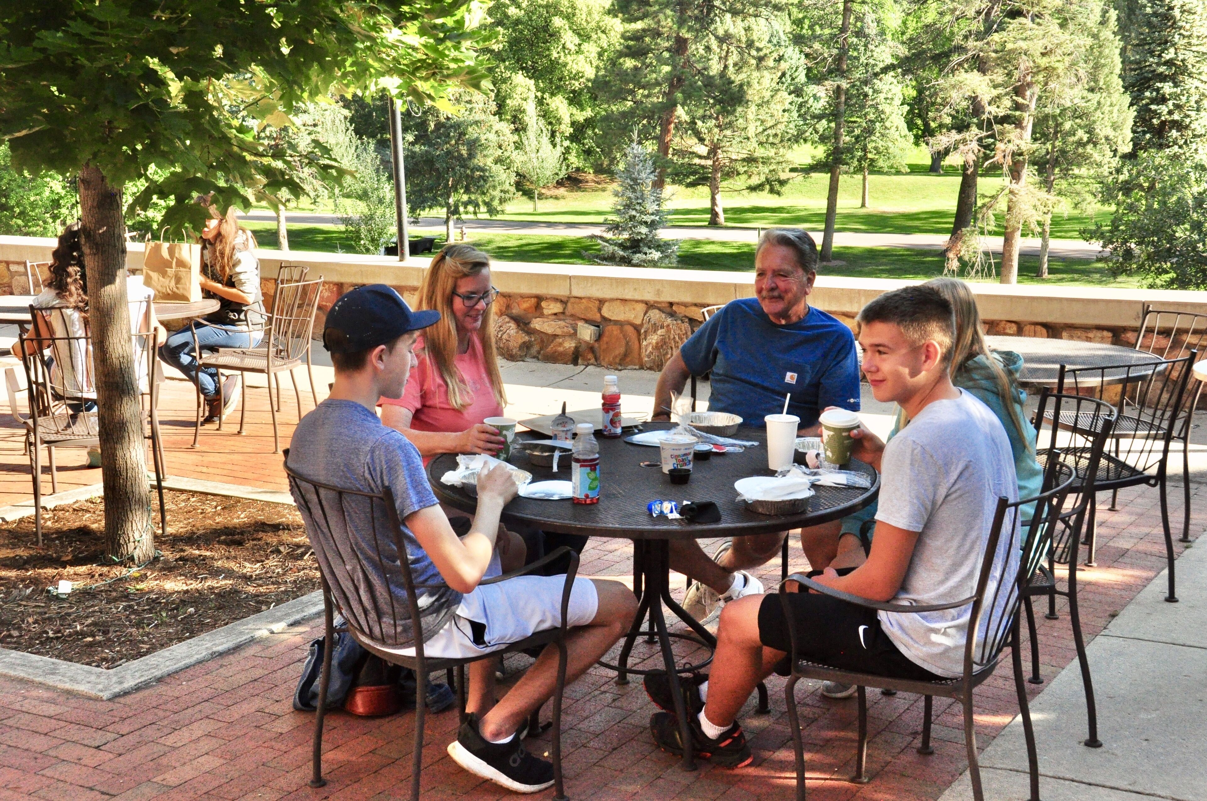 A group of people seated around a round table outdoors, with a stone wall and trees in the background.