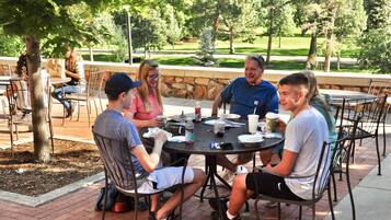 A group of people seated around a round table outdoors, with a stone wall and trees in the background.