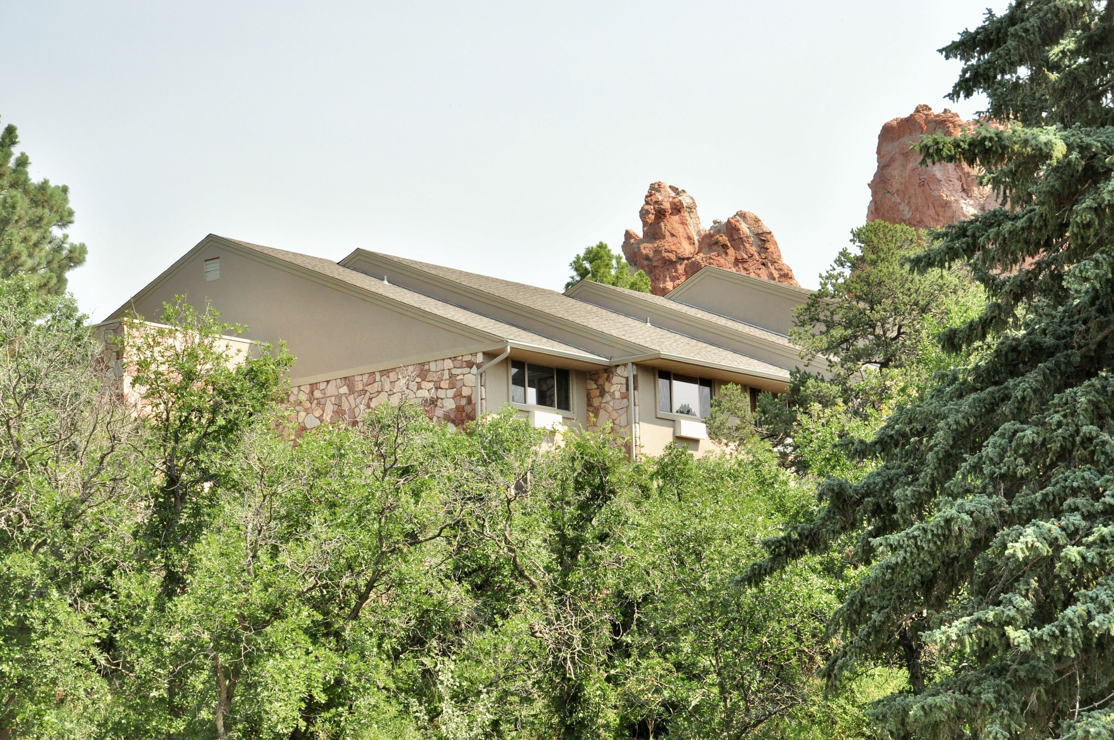 A two-story building with a stone facade, surrounded by dense green trees and a prominent rock formation in the background.