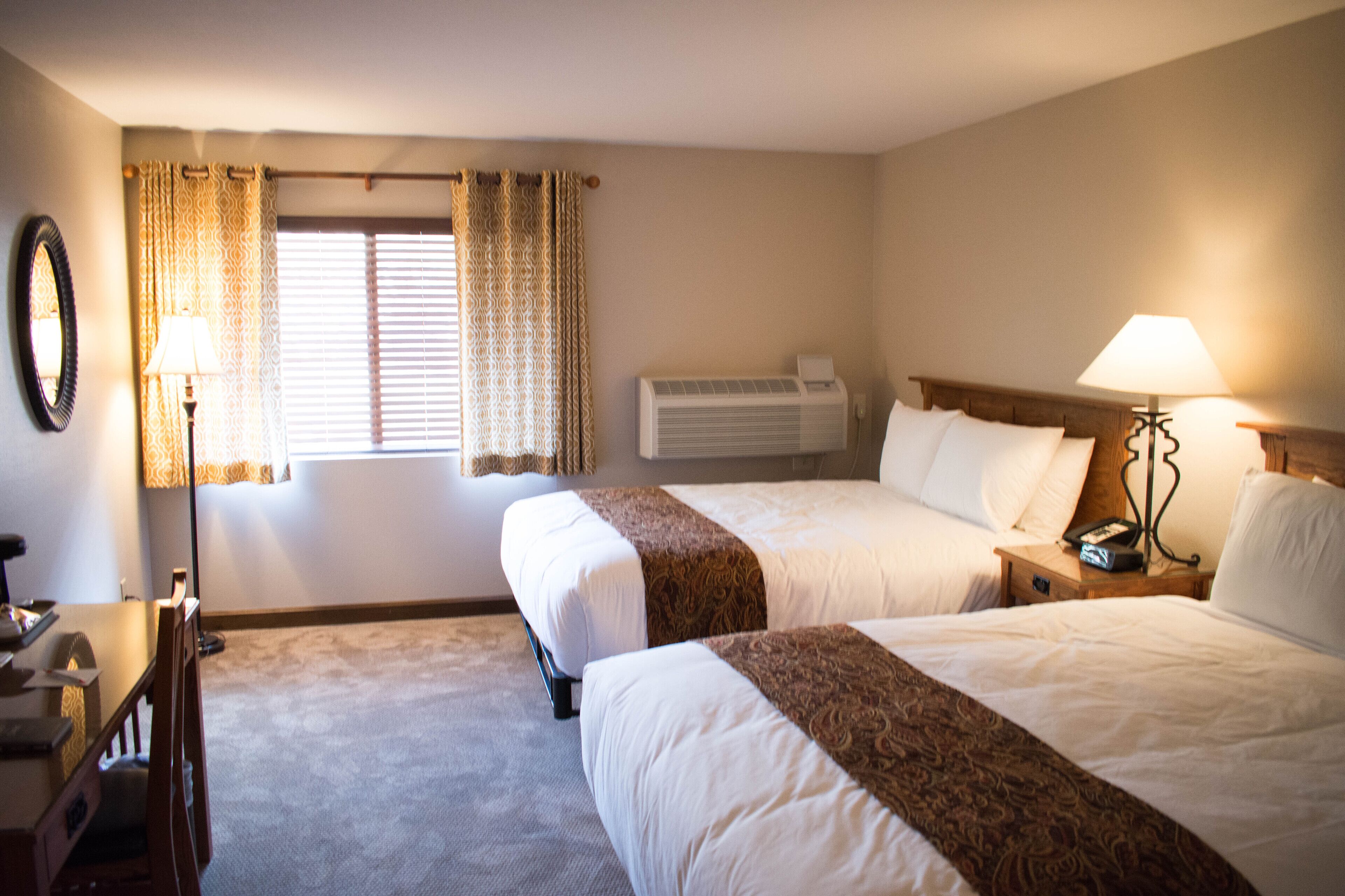 A hotel room with two beds, a desk, a chair, a lamp, a mirror, and an air conditioning unit.