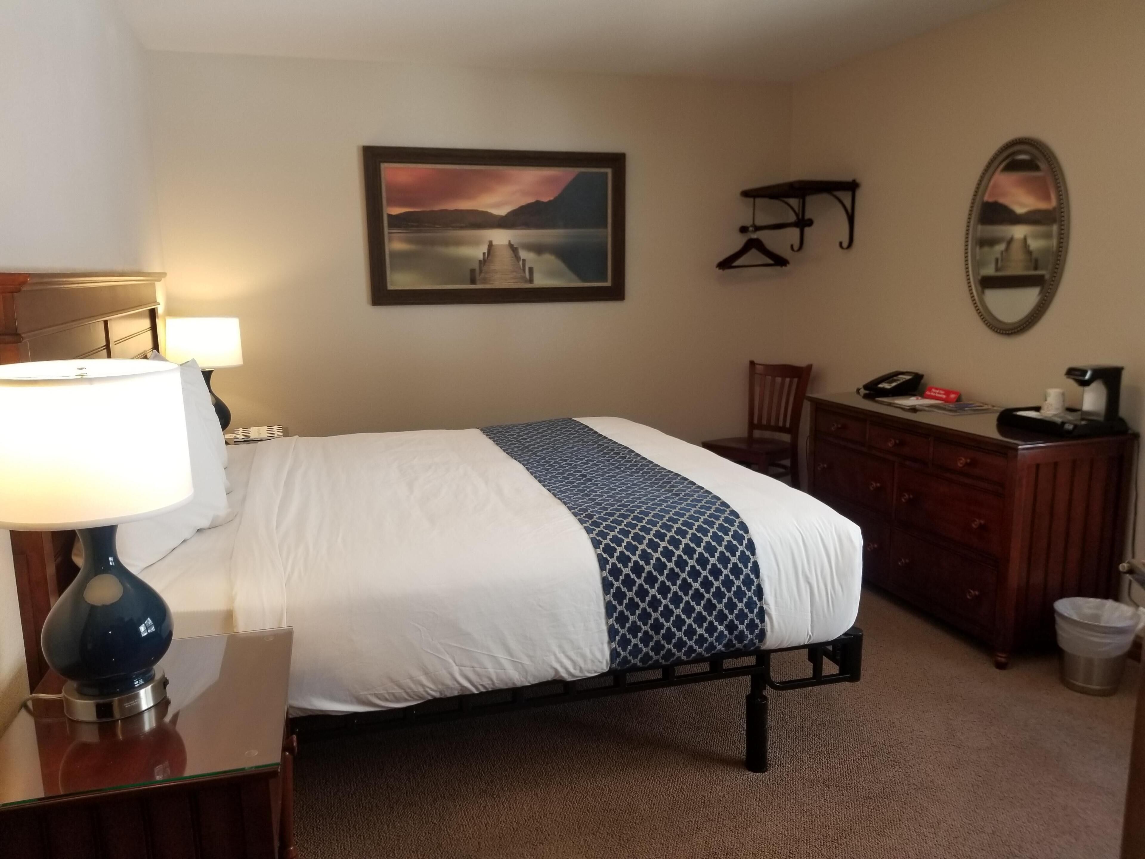 Une chambre d’hôtel comprenant un lit, un bureau avec une lampe, une commode, une chaise, une lampe de bureau, un tableau encadré au mur, un miroir et une poubelle.