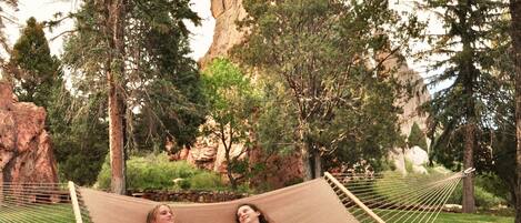 Two people relaxing on a hammock in a grassy area with trees and a rock formation in the background.