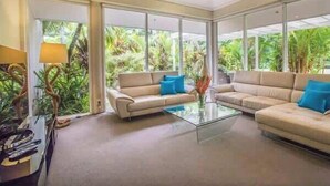 A spacious living room with two beige sofas, a glass coffee table, and a large window offering a view of lush greenery. - Port Douglas Mirage Villa 408 (Port Douglas)