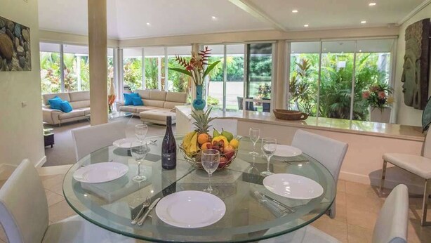 A dining area with a round glass table set for six, surrounded by white chairs. There is a fruit bowl on the table and a vase with flowers. The room has large windows with a view of greenery outside. - Port Douglas Mirage Villa 408 (Port Douglas)