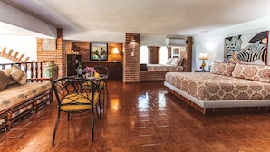 A spacious room with a brick fireplace, a bed with patterned bedding, a dining area with a table and chairs, and a large window.
