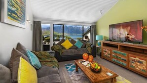 Un salon avec un canapé, une table basse et un téléviseur. Il y a une fenêtre donnant sur un paysage de montagnes et d’un lac.