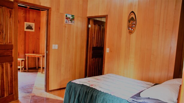 Una habitación de madera con una cama, una silla y una puerta que da a otra estancia.