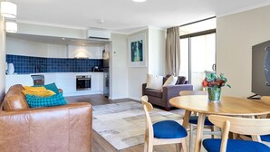 Un salon moderne avec un canapé en cuir marron, une table à manger en bois et des chaises bleues, une cuisine en arrière-plan et une grande fenêtre avec des rideaux.