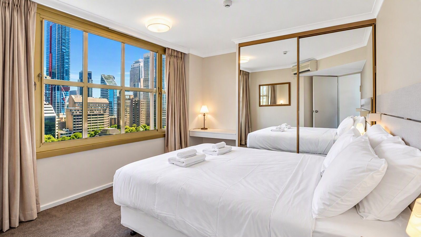 A hotel room with a large window, a bed, a bedside table with a lamp, and a mirror.