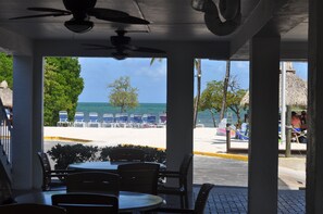 An outdoor seating area with tables and chairs, overlooking a beach with lounge chairs and a thatched-roof structure. - Oceanfront, 2 Bedroom Unit At Ocean Pointe Condominium (Tavernier)