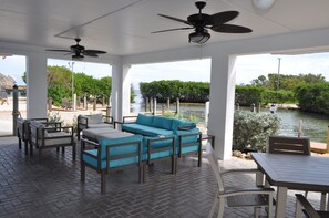 A covered outdoor seating area with a sofa and chairs, a table, and a view of a water body with trees. - Oceanfront, 2 Bedroom Unit At Ocean Pointe Condominium (Tavernier)