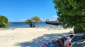 A beach with lounge chairs, a thatched-roof structure, and a pier in the background. - Oceanfront, 2 Bedroom Unit At Ocean Pointe Condominium (Tavernier)