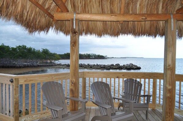 A wooden deck with three Adirondack chairs facing a body of water, a thatched roof structure, and a rocky shoreline in the background.