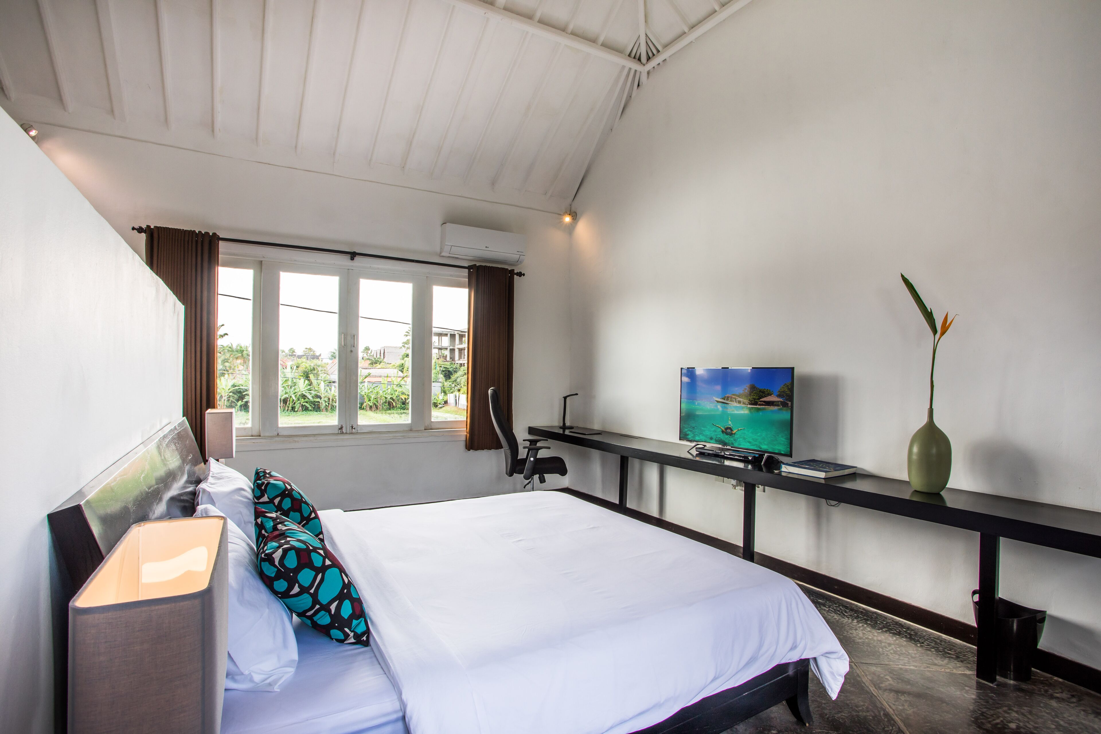 Ein Hotelzimmer mit einem großen Bett, einem Schreibtisch mit Fernseher, einer Vase und einem Fenster mit Vorhängen.