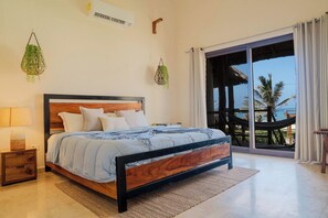 A bedroom with a bed, bedside table, lamp, hanging plants, and a view of the outdoors.