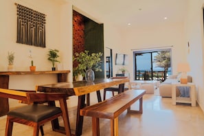 A modern living room with a wooden dining table, benches, and a sofa. There are potted plants and a large window with a view.