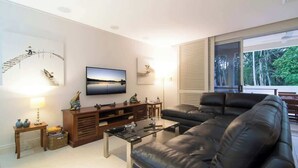 A modern living room with a large black leather sofa, a wooden entertainment unit, a flat-screen TV, and a large window with blinds.